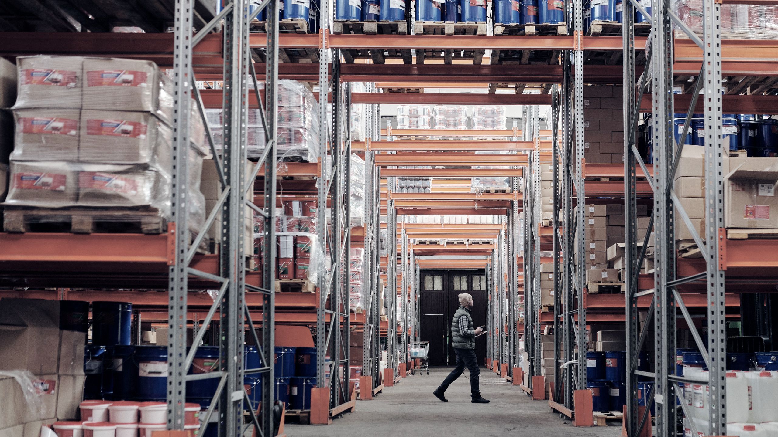 Almacén de una distribuidora: pasillo entre racks con tarimas cargadas y un trabajador revisando el inventario.