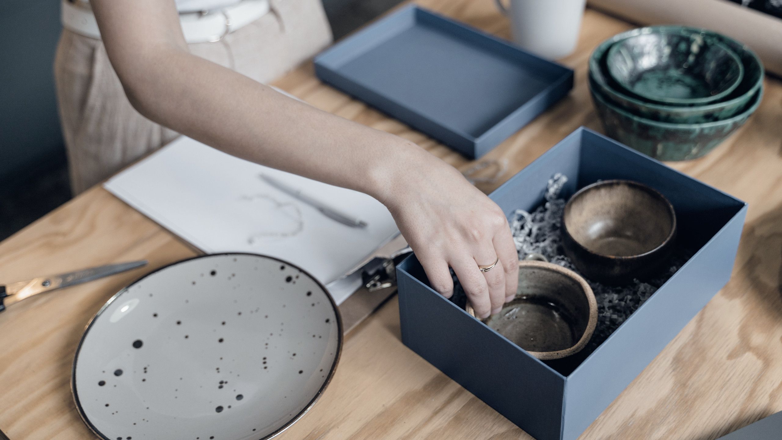 Preparación de un pedido de e-commerce: unas manos acomodan tazas de cerámica en una caja con relleno de papel.