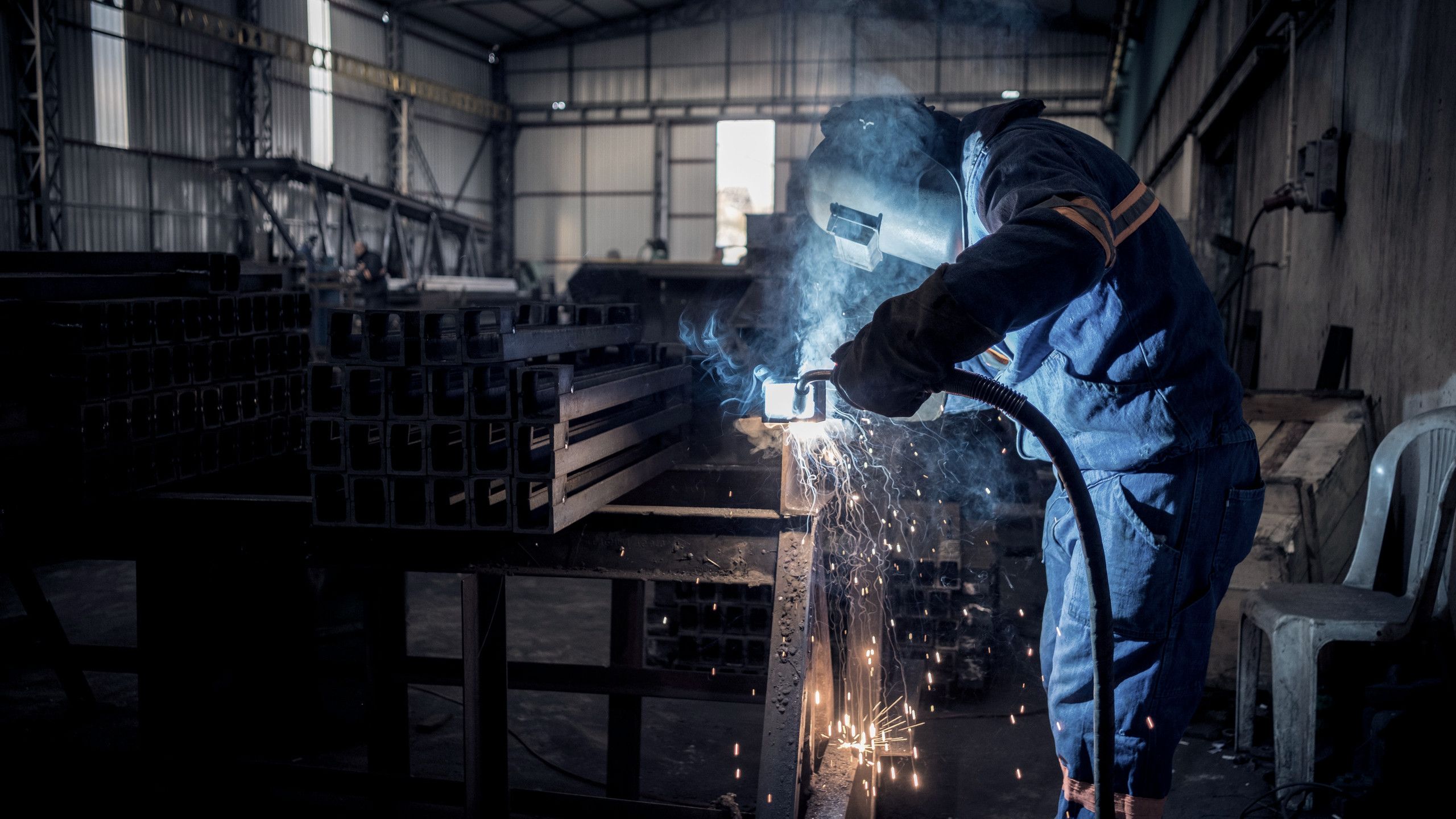 Planta de manufactura metalmecánica: un soldador con careta y overol trabaja una pieza de acero.
